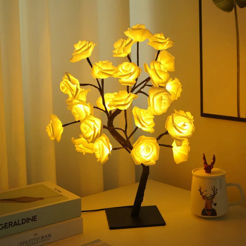 Warm yellow LED rose flower lamp on a black tree-like stand, illuminating a cozy room with a soft glow. Positioned on a table with decorative items including a reindeer mug and stacked books, creating a relaxing and elegant ambiance. Perfect for romantic home decor and ambient lighting.