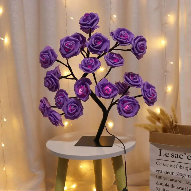 Purple LED rose flower lamp with a black tree-like base, illuminating a cozy room. Placed on a small white table, the vibrant purple roses glow warmly, creating a soft and romantic ambiance. String lights hang in the background, enhancing the aesthetic. Ideal for decorative lighting and unique home decor.