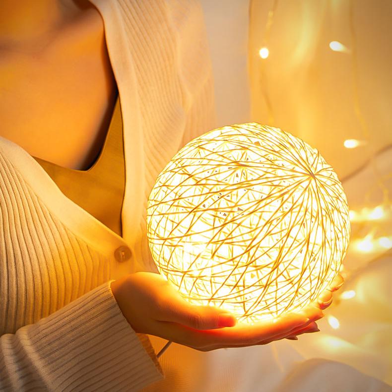 Warm rattan ball lamp held in hands, emitting a soft, golden glow that creates a cozy, comforting ambiance. The woven texture diffuses the light beautifully, ideal for adding warmth to any room. Soft fairy lights in the background enhance the serene, inviting atmosphere, perfect for relaxation or meditation spaces.
