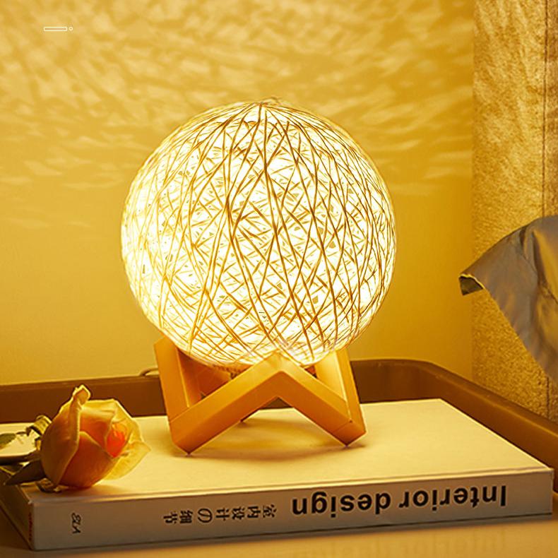 Elegant woven rattan ball lamp emitting a warm, cozy glow, perfect for ambient lighting in bedrooms or living spaces. The lamp sits on a wooden stand, creating beautiful shadow patterns on the surrounding surfaces. Styled on a table with a rose and a design book, this lamp adds a natural and relaxing touch to any decor.