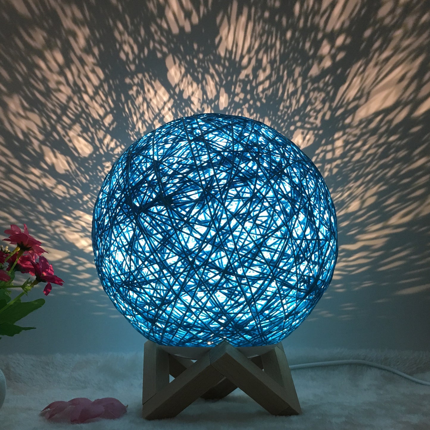 Blue rattan ball lamp on a wooden stand, casting intricate shadow patterns across the wall, creating a serene and artistic ambiance. The woven design diffuses the light softly, producing a calming glow. Accented with pink flowers, this lamp is ideal for adding a unique touch to bedrooms, living rooms, or relaxation spaces.