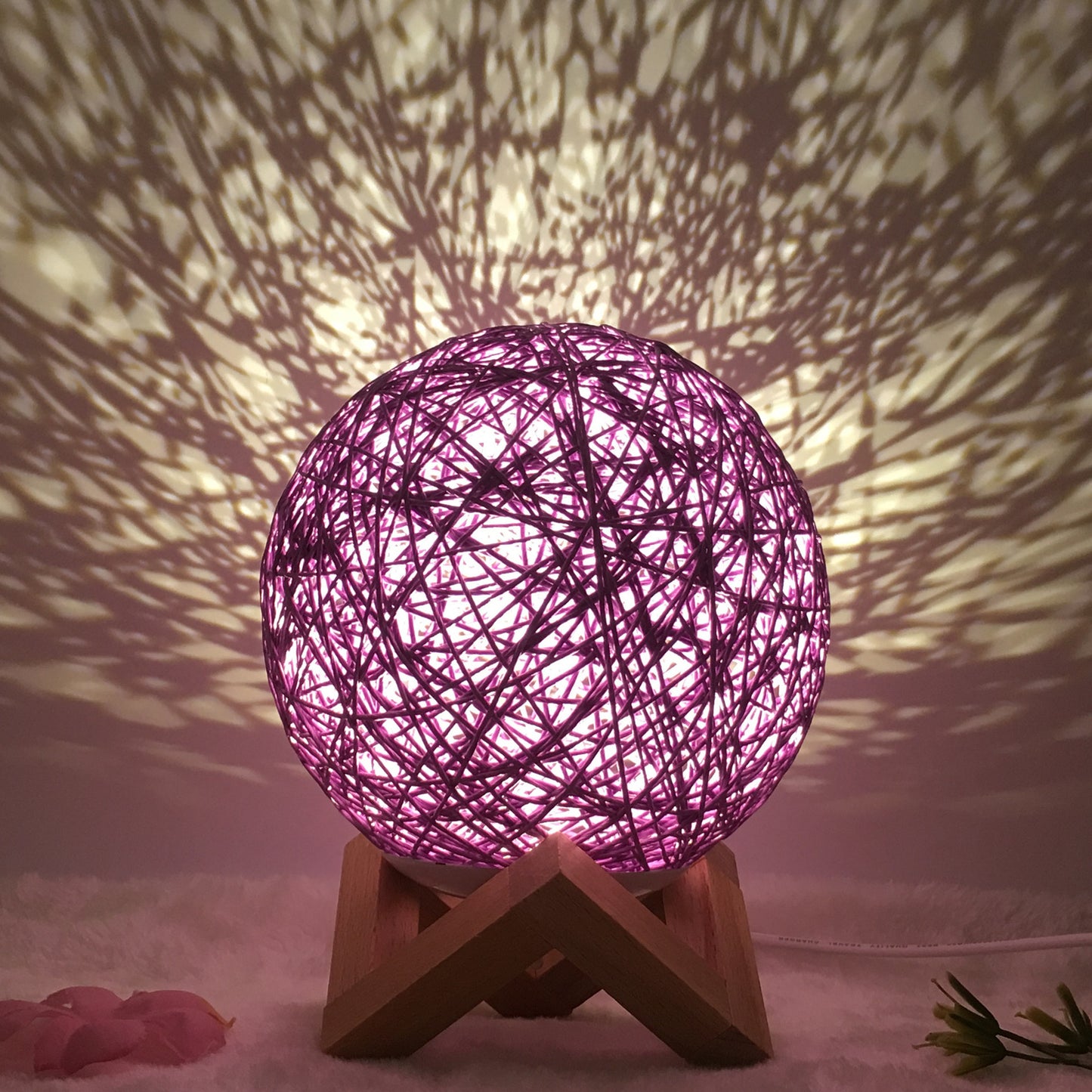 Purple rattan ball lamp on a wooden stand, casting intricate shadow patterns across the wall, creating a unique and cozy atmosphere. The woven design diffuses light softly, making it ideal for relaxing spaces such as bedrooms or living rooms. Accented with delicate floral decor, this lamp adds a touch of warmth and elegance to any setting.