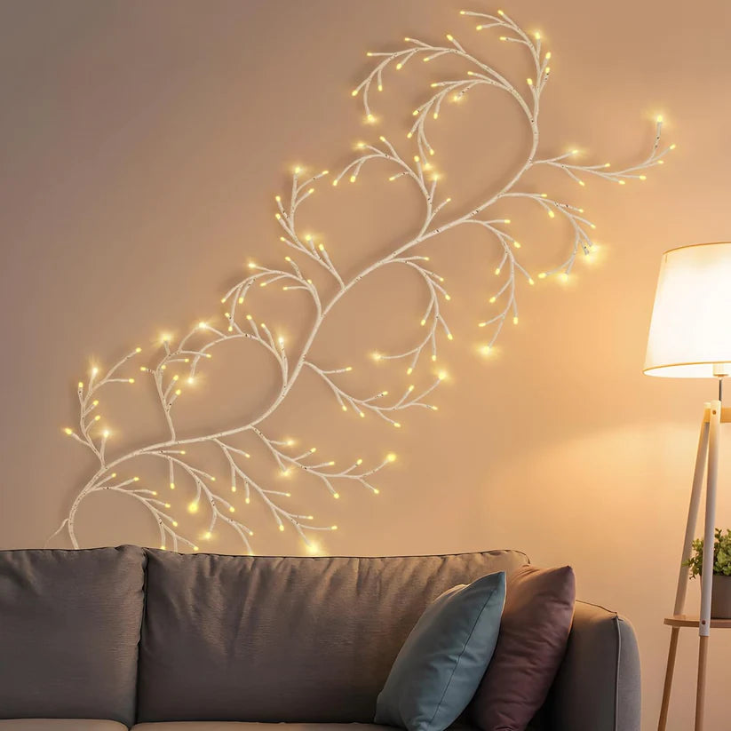 This image displays a cozy and artistic living room setup featuring a unique wall decoration of illuminated branches. The branches, adorned with warm, glowing LED lights, mimic the appearance of tree limbs spreading organically across the wall, creating a soft and inviting ambiance. This nature-inspired decor adds a whimsical, yet refined touch to the room, perfectly complementing the neutral tones of the sofa and cushions. A modern floor lamp to the right adds an additional layer of soft lighting.