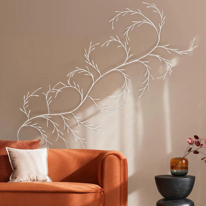 a beautiful wall decor element in the form of a branch-like structure that creates an organic, flowing design across the wall. The structure has delicate, twig-like branches that gently curve and meander, giving a subtle sense of movement and adding a natural, artistic touch to the space. The decor is complemented by a warm, burnt-orange sofa and a sleek, modern side table with a small vase, creating an elegant yet cozy ambiance. This setup would suit a minimalist or nature-inspired interior design style.