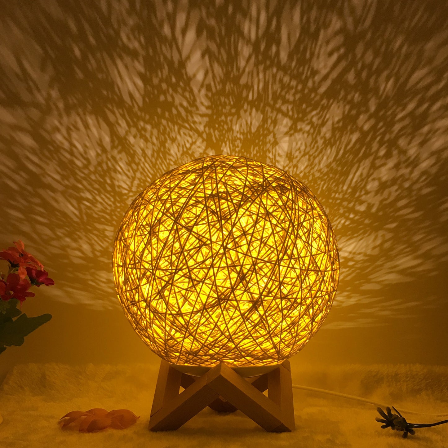 Golden rattan ball lamp on a wooden stand, emitting a warm, radiant glow and casting intricate shadow patterns on the wall. The woven design creates a cozy and artistic ambiance, ideal for bedrooms or living rooms. Accented with subtle floral decor, this lamp brings a natural, inviting warmth to any space.