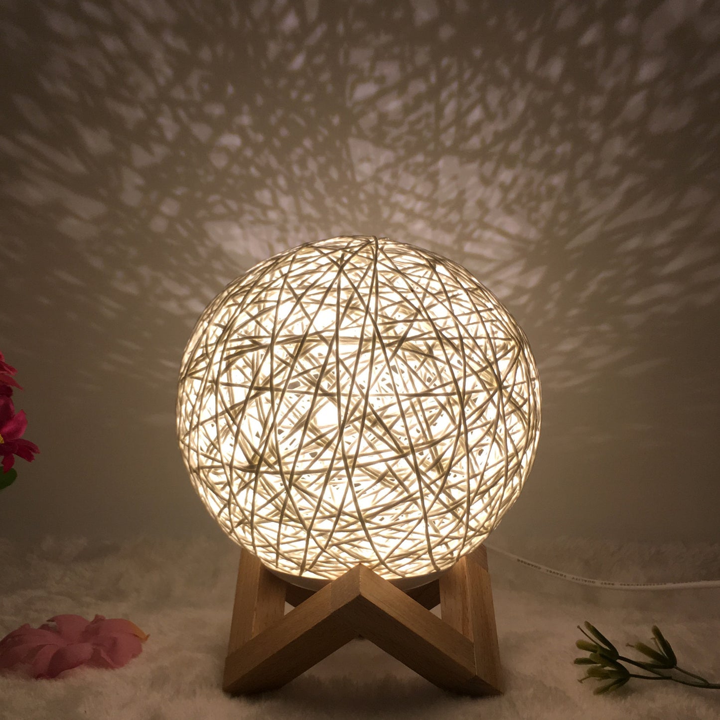 Warm rattan ball lamp on a wooden stand, projecting intricate shadow patterns on the wall, creating a cozy and artistic ambiance. The woven design of the lamp diffuses light softly, perfect for enhancing relaxation in bedrooms or living areas. Surrounded by delicate floral accents, this lamp adds a touch of natural elegance to any space.