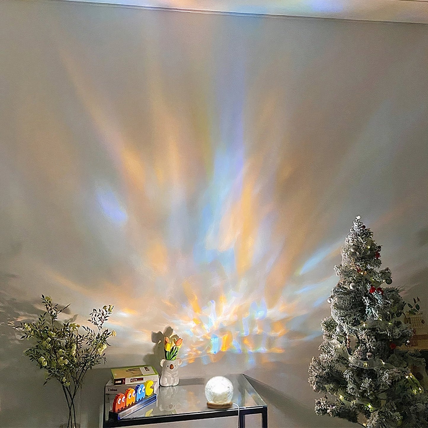 This image features a decorative lamp on a tabletop, casting beautiful, multi-colored light patterns across a plain wall. The light radiates in soft rainbow hues, creating a calming and atmospheric effect. The setup includes minimal decor, with a small Christmas tree in the corner and a potted plant, adding a touch of nature to the scene. This lighting effect brings a festive, cozy ambiance, perfect for holiday or relaxation settings.