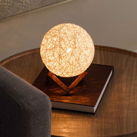 Minimalist rattan ball lamp on a wooden stand, casting a soft, warm light perfect for ambient or accent lighting in cozy spaces. Positioned on a dark coffee table with a closed book beneath it, this lamp adds a touch of natural elegance to modern interiors.