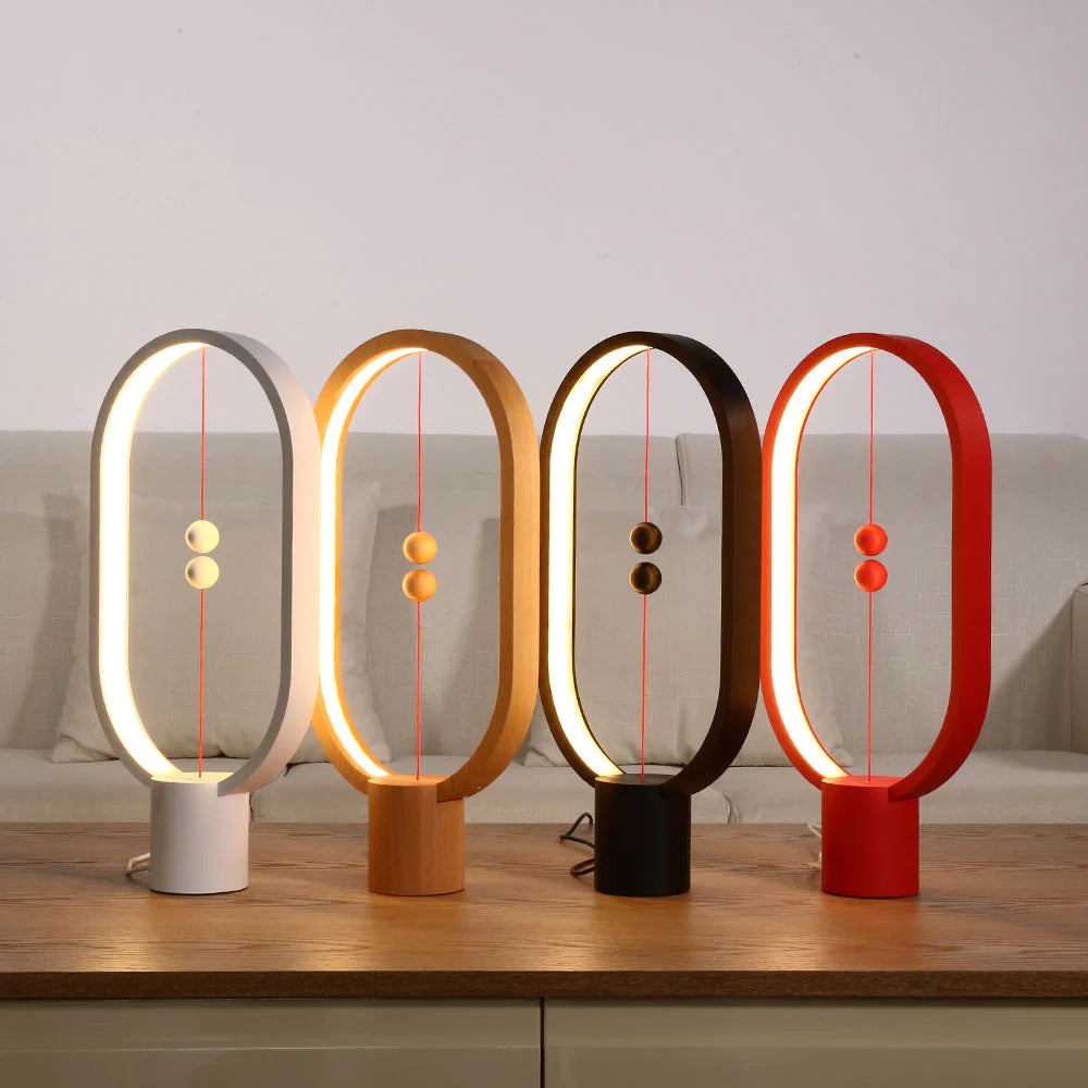 A set of four minimalist magnetic balance lamps in varying finishes — white, natural wood, black, and red — each featuring an oval frame with a warm LED light strip along the inner edge. Positioned on a wooden table against a neutral backdrop, these lamps create a cohesive yet vibrant display. Each lamp has two magnetic spheres suspended on a red cord, floating in the center when aligned to switch on the light. This stylish collection showcases modern design, adding both functional lighting and an artistic