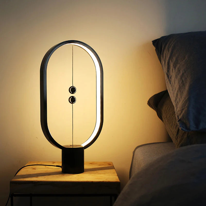 A sleek, modern magnetic balance lamp emits a warm, ambient glow beside a bed, creating a cozy nighttime atmosphere. The lamp’s minimalist oval frame houses a subtle light strip along its inner edge, while two magnetic balls are suspended in the center on a thin cord, acting as the switch mechanism. Placed on a wooden bedside table, the lamp's soft lighting provides a calming accent ideal for a restful setting, complementing the cozy bedding beside it.