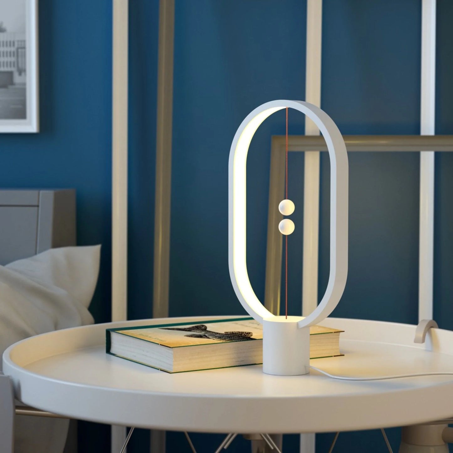 A contemporary magnetic balance lamp with a sleek white frame sits gracefully on a round white table, its soft light casting a warm glow. The design features a minimalistic oval shape with an illuminated inner edge, highlighted by two suspended magnetic balls in the center that function as a unique on-off switch. Positioned alongside a closed book, the lamp adds a touch of modern elegance and tranquility to the room, complemented by a deep blue wall backdrop. The setup creates a serene reading or relaxation