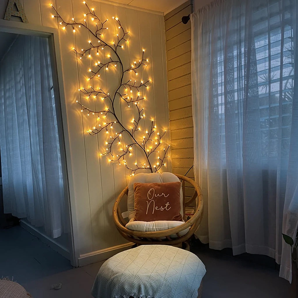 Cozy reading nook with warm LED branch lights on wall, soft armchair, and 'Our Nest' pillow. Ambient lighting creates a peaceful, inviting corner ideal for relaxation. Perfect decor for a hygge-inspired space or nature-themed interiors, enhancing home aesthetics with ambient lighting in a comfortable seating area.