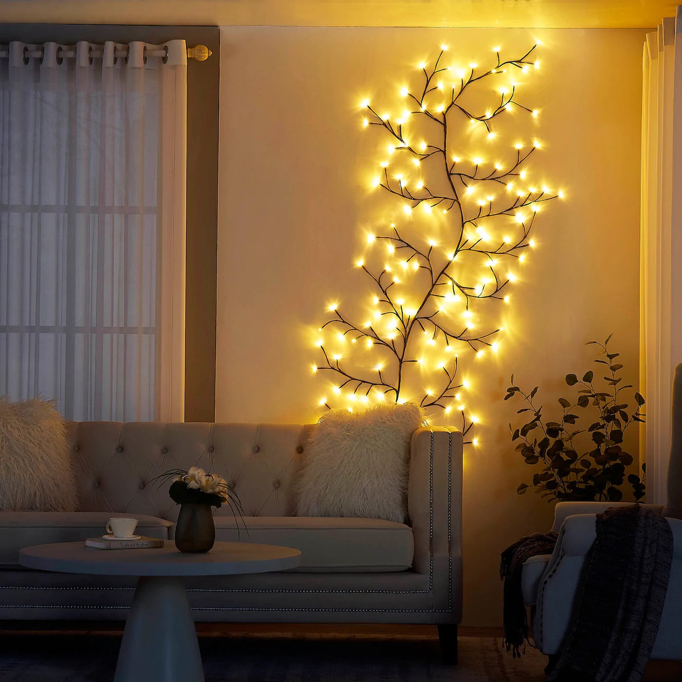 Elegant living room with wall-mounted LED branch lights, creating a warm, ambient glow. Cozy atmosphere enhanced by tufted sofa with fluffy pillows, round coffee table, and natural greenery. Ideal lighting decor for modern and minimalist interiors, adding a touch of nature-inspired illumination.