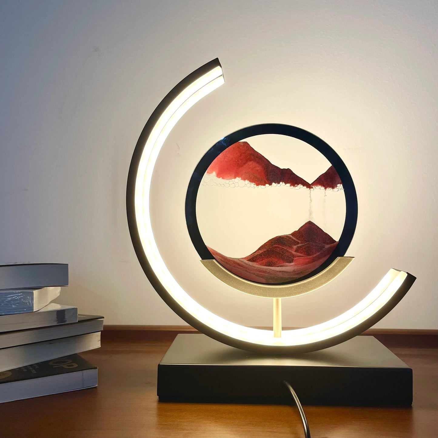 A crescent LED sand art lamp with vibrant red sand creating a mountain-like landscape, enclosed in a circular frame. This modern piece adds a soothing visual to any desk or shelf, perfect for a unique ambient lighting experience.