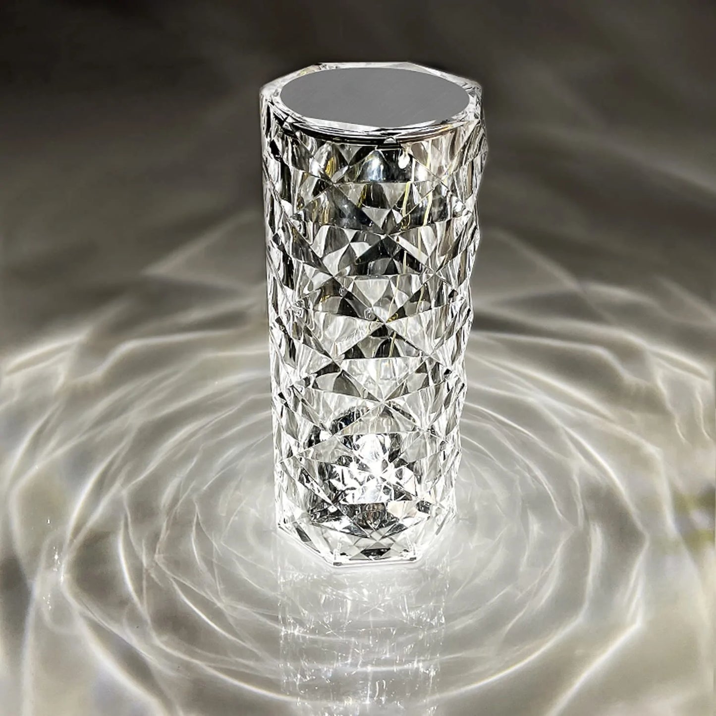 A crystal LED lamp with a tall, diamond-cut design, casting mesmerizing light patterns in concentric circles around it. The faceted surface of the lamp reflects light beautifully, creating a sophisticated ambiance with an almost water-like ripple effect. This elegant, modern lamp adds a touch of luxury and visual interest to any space, enhancing the room's decor with its unique and eye-catching lighting.