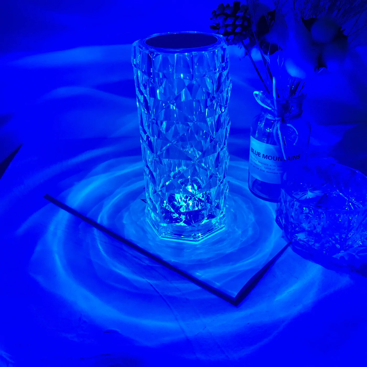 A crystal LED lamp with a hexagonal, diamond-cut design illuminated in a vibrant blue hue. The faceted crystal surface reflects light, creating captivating, ripple-like patterns that spread outwards, adding a soothing ambiance to the space. Perfect as a statement piece for modern decor, this lamp’s unique shape and striking blue illumination bring an artistic and luxurious touch to any room.