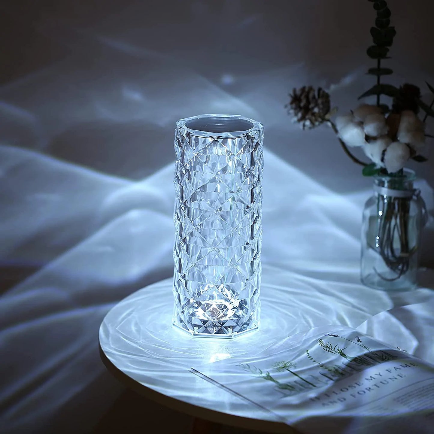 A tall, diamond-patterned crystal LED lamp illuminating a soft, cool white light. The intricate crystal design casts delicate, wave-like reflections on the surrounding surfaces, creating a serene, watery ambiance. This sophisticated lamp is positioned on a white table, with a minimalist decor setting that includes a glass vase and a small floral arrangement. Ideal for adding a touch of elegance and calming light to a modern living space.