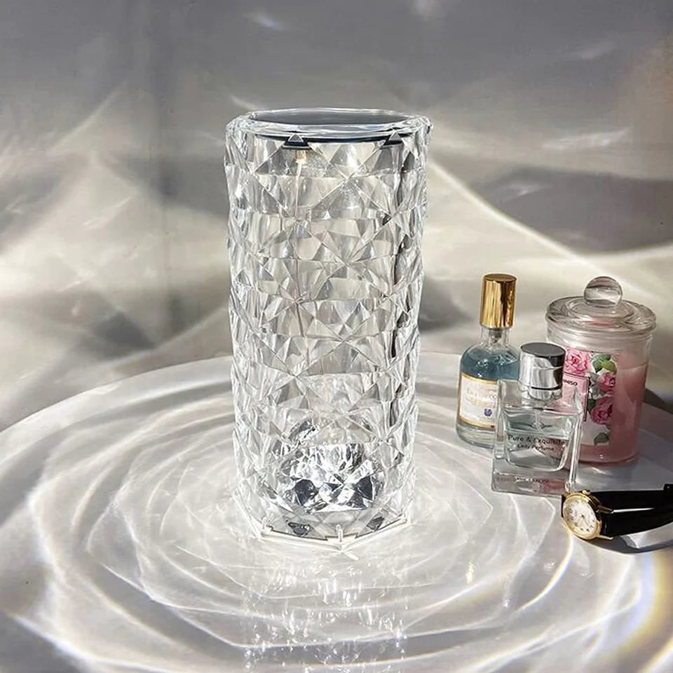 A tall, crystal LED lamp with a diamond-cut pattern rests on a reflective surface, creating intricate, shimmering light effects that cascade outward. Surrounding the lamp are small decorative items, including perfume bottles and a candle jar, adding a touch of elegance and luxury to the setting. The lamp’s clear, faceted design enhances the ambient lighting, making it a sophisticated choice for accentuating any room decor.