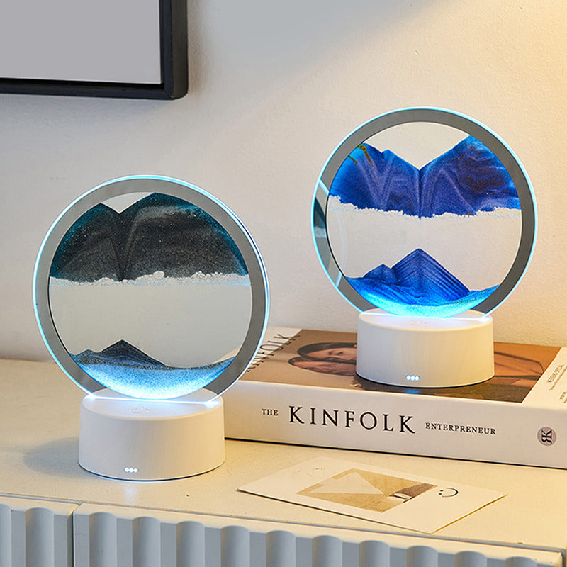 Two round LED sand art lamps with dynamic sandscapes in contrasting black and blue hues, each creating serene mountainous formations. Set on a white base, the lamps emit a gentle ambient glow, adding a calming atmosphere to any interior. Placed on a tabletop near a "Kinfolk Entrepreneur" book, perfect for modern, minimalist decor.