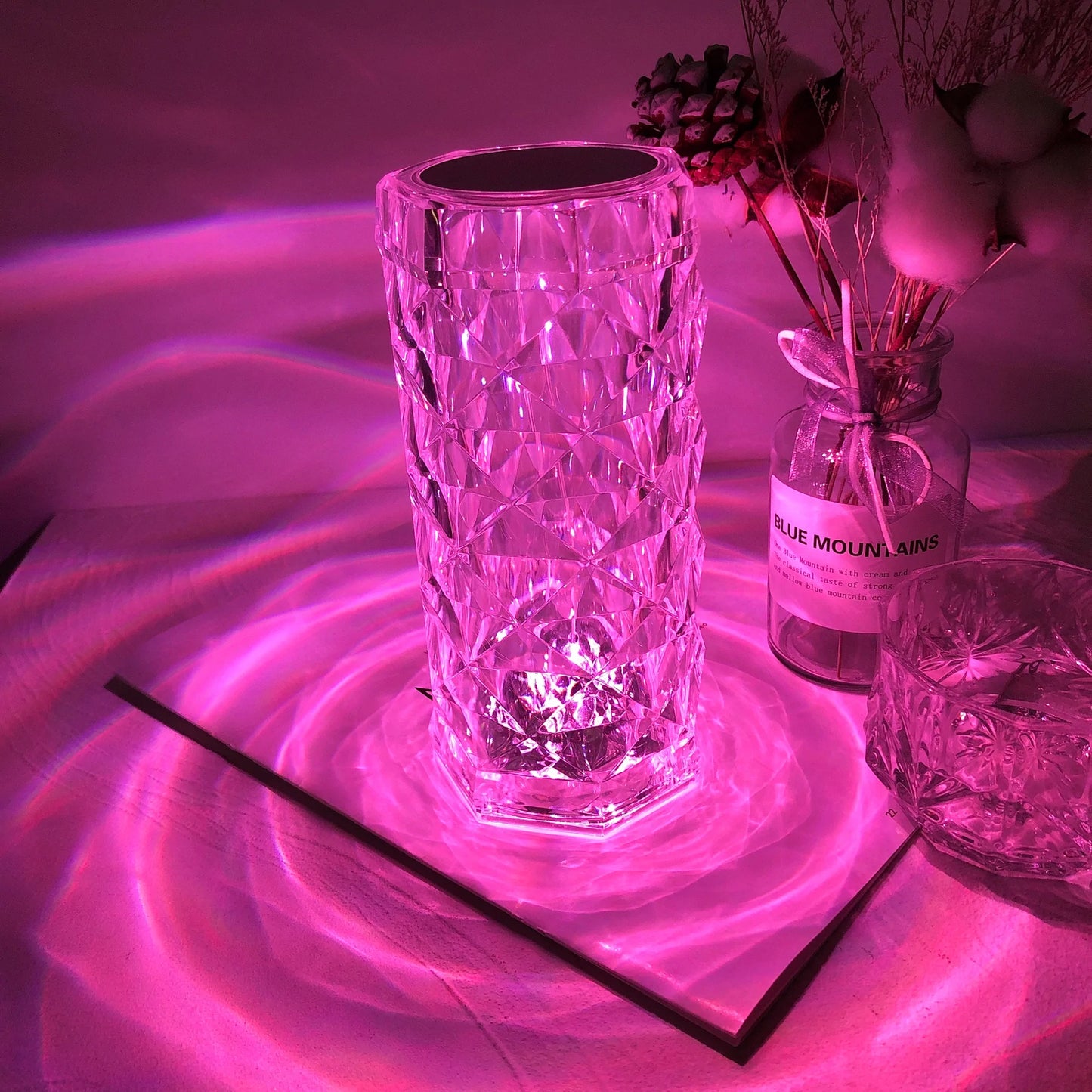 A tall, diamond-patterned crystal LED lamp glowing with a vibrant pink light. The faceted crystal design creates mesmerizing, ripple-like reflections on the surface around it, casting a warm and enchanting ambiance. Accompanied by decorative elements, including a glass bottle labeled "Blue Mountains" and a smaller crystal glass, this lamp adds a luxurious and romantic atmosphere, perfect for setting a cozy and stylish mood in any room.