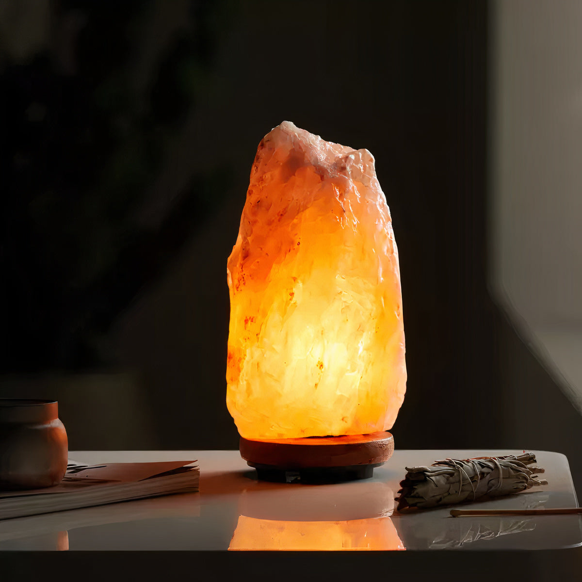 Warmly glowing Himalayan salt lamp with a rugged, crystalline texture, radiating a soothing amber light. It stands on a small wooden base on a white tabletop, accompanied by a bundle of sage and a small, minimalistic clay pot, creating a cozy and serene ambiance.