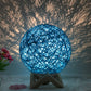 Blue rattan ball lamp on a wooden stand, casting intricate shadow patterns across the wall, creating a serene and artistic ambiance. The woven design diffuses the light softly, producing a calming glow. Accented with pink flowers, this lamp is ideal for adding a unique touch to bedrooms, living rooms, or relaxation spaces.