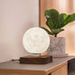 A levitating moon lamp with a realistic lunar surface, softly glowing above a wooden base on a modern sideboard. Positioned near a green potted plant, it enhances the minimalist decor, adding a calming, celestial touch to the room.