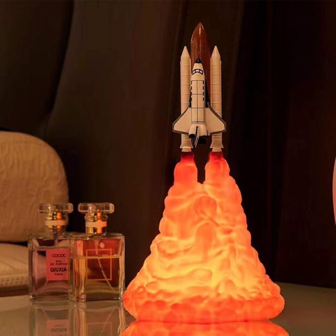 Illuminated rocket lamp designed as a space shuttle blasting off, with vibrant LED flames in a warm orange glow, creating a liftoff effect on a bedside table. Positioned beside two luxury perfume bottles, this unique lamp adds a touch of adventure and a modern aesthetic to the room, perfect as decor for space enthusiasts and kids’ bedrooms.