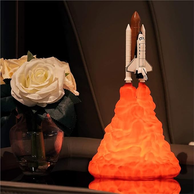 Space shuttle rocket lamp with LED flame effect next to a white rose in a glass vase on a reflective surface. The rocket lamp emits a vibrant orange glow from its base, simulating a realistic launch thrust effect, making it an ideal piece for space-themed decor or as a unique ambient night light. The soft lighting and realistic details of the shuttle create a dramatic effect, perfect for adding a touch of adventure and fascination to a room.