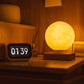 A glowing levitating moon lamp emits a soft, warm light on a wooden bedside table, creating a cozy, ambient atmosphere. Positioned next to a digital clock reading "01:39," the lamp illuminates the room with its realistic lunar surface texture, enhancing the serene nighttime setting. Perfect for a calming, celestial-inspired decor.