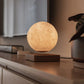 A levitating moon lamp emitting a warm, golden glow, floating above a minimalist wooden base on a sleek console. Positioned in a cozy setting, it adds a soft, ambient light, creating a tranquil atmosphere perfect for relaxation. The realistic lunar surface texture adds a touch of celestial elegance to the room.