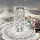A tall, crystal LED lamp with a diamond-cut pattern rests on a reflective surface, creating intricate, shimmering light effects that cascade outward. Surrounding the lamp are small decorative items, including perfume bottles and a candle jar, adding a touch of elegance and luxury to the setting. The lamp’s clear, faceted design enhances the ambient lighting, making it a sophisticated choice for accentuating any room decor.