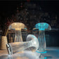 Three crystal mushroom-shaped LED lamps are placed on a surface against a cityscape background, with two standing upright and one lying on its side. The lamps emit a soft, refracted glow in warm and cool tones, casting intricate patterns of light and shadow. Their ribbed, transparent design enhances the luminous effect, making these lamps both a decorative and functional piece suitable for modern and contemporary settings.