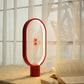 A red magnetic balance lamp with a sleek, oval-shaped frame, casting a warm glow on a beige tabletop. The lamp features two small magnetic spheres, also in red, that suspend in the center when aligned to activate the light. Positioned in front of a softly lit window with sheer curtains, this lamp creates a cozy and ambient atmosphere, ideal for reading or unwinding. An open book lies next to the lamp, enhancing the scene’s inviting and relaxed setting. This piece adds a modern yet vibrant accent to any spac