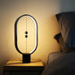 A sleek, modern magnetic balance lamp emits a warm, ambient glow beside a bed, creating a cozy nighttime atmosphere. The lamp’s minimalist oval frame houses a subtle light strip along its inner edge, while two magnetic balls are suspended in the center on a thin cord, acting as the switch mechanism. Placed on a wooden bedside table, the lamp's soft lighting provides a calming accent ideal for a restful setting, complementing the cozy bedding beside it.