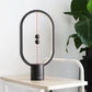 A modern magnetic balance lamp with an oval frame and minimalistic design, set on a light wooden table with a leafy plant in the background. The lamp has two spherical magnetic balls suspended on a red string within the frame. When the balls are aligned in the center, the lamp illuminates, creating a warm, ambient glow. Its black matte finish contrasts subtly with the light table and white background, adding a sophisticated and contemporary touch to any interior space. This piece combines art and functional