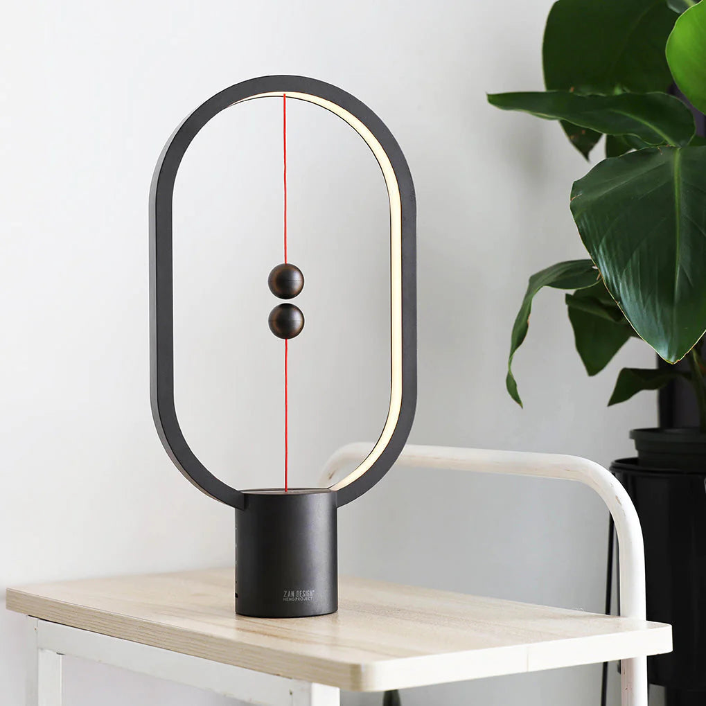 A modern magnetic balance lamp with an oval frame and minimalistic design, set on a light wooden table with a leafy plant in the background. The lamp has two spherical magnetic balls suspended on a red string within the frame. When the balls are aligned in the center, the lamp illuminates, creating a warm, ambient glow. Its black matte finish contrasts subtly with the light table and white background, adding a sophisticated and contemporary touch to any interior space. This piece combines art and functional