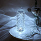 A tall, diamond-patterned crystal LED lamp illuminating a soft, cool white light. The intricate crystal design casts delicate, wave-like reflections on the surrounding surfaces, creating a serene, watery ambiance. This sophisticated lamp is positioned on a white table, with a minimalist decor setting that includes a glass vase and a small floral arrangement. Ideal for adding a touch of elegance and calming light to a modern living space.