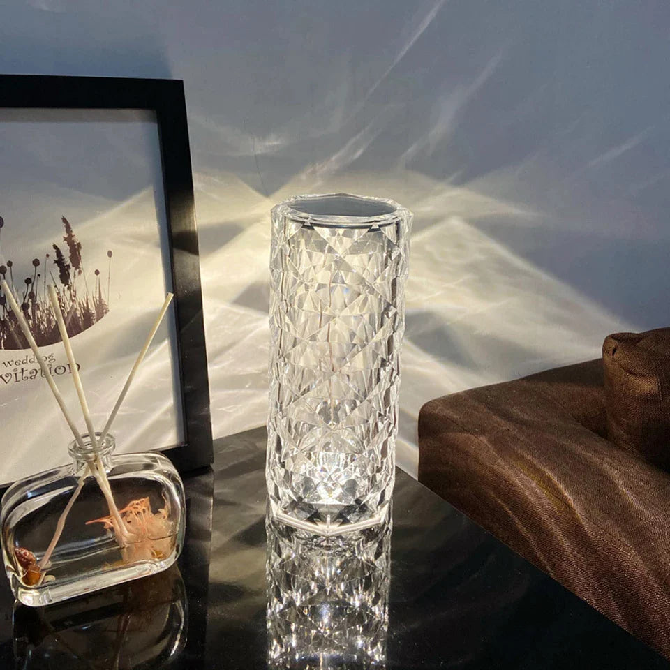 A crystal LED lamp with a diamond-cut design sits elegantly on a dark surface, casting intricate light patterns on the wall. Its clear, faceted surface creates a shimmering effect, adding a luxurious ambiance to the room. Nearby, a delicate reed diffuser and framed artwork enhance the stylish, serene decor, making this a perfect choice for modern, ambient lighting.