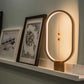 A minimalist wooden magnetic balance lamp sits elegantly on a picture ledge, casting a warm glow that enhances the surrounding artwork. The lamp’s oval frame is illuminated along its inner edge, creating a soft ambiance that accentuates the wall art. The distinctive design features two small magnetic spheres suspended on a red cord, floating in the center when activated. This piece combines modern aesthetics with functional lighting, making it a stylish addition to any artful space.